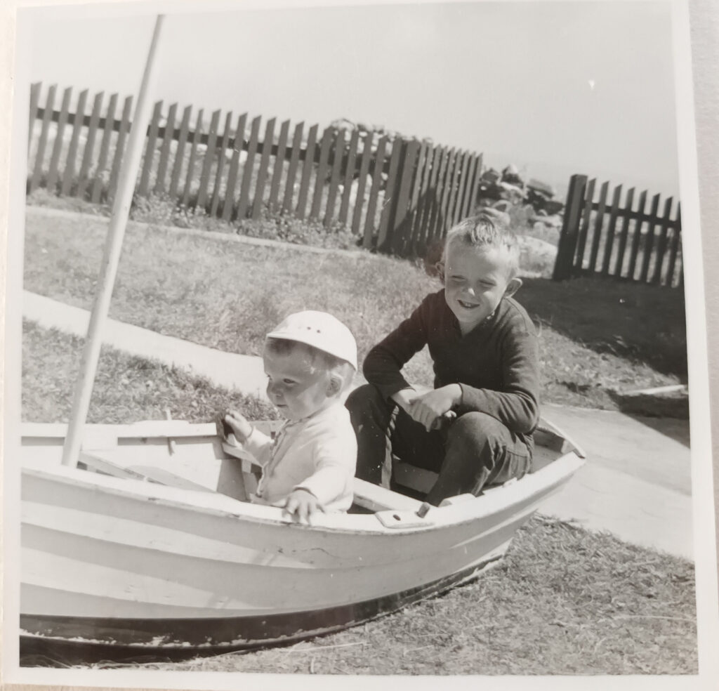 Svartvitt foto av två barn som sitter i en liten träbåt på land. Staketet i bakgrunden har en öppen grind.