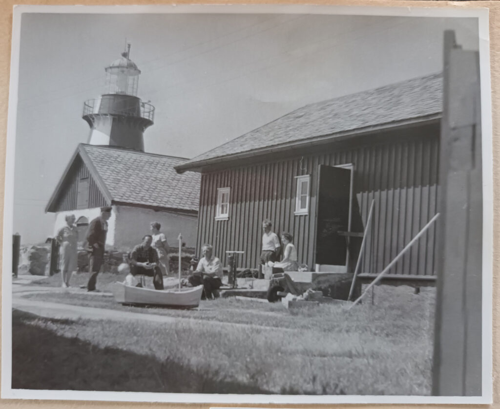 Svart-vitt foto med ett sällskap som står och sitter inne på gårdstomten. De är lätt klädda i vardagskläder. Framför sällskapet står en liten båt med ett barn i. Fyrtornet sticker upp i bakom en byggnad.