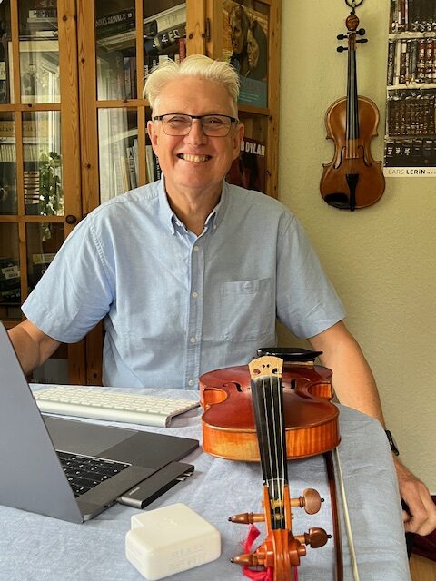Porträttfoto: Göran Premberg sitter vid ett bord med en dator och fiol på. Han har vitt hår och ler brett mot kameran. 