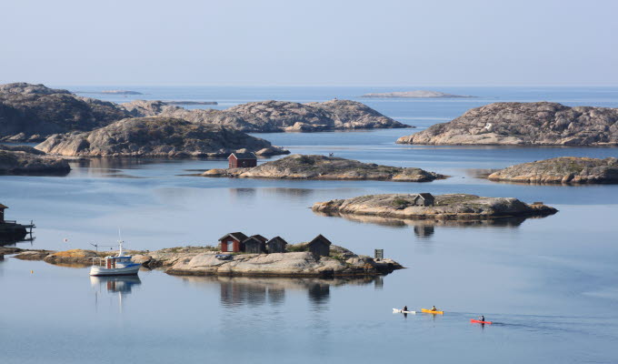 Foto. En havsvy. Ett antal stenklippor reser sig från havet, i förgrunden syns en vit båt och tre paddlare.