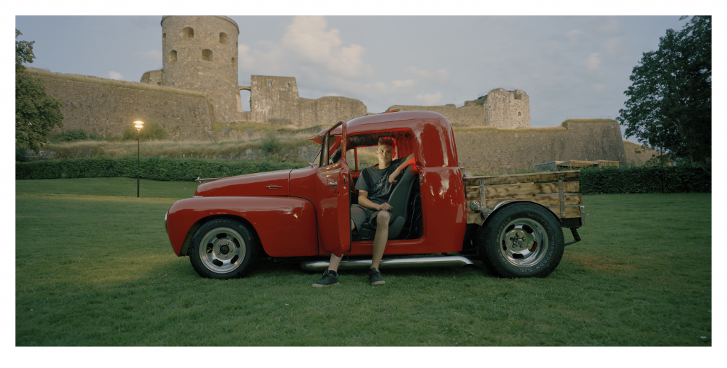 Foto. En ung man sitter i en glänsande röd EPA med 50-tals front står framför en medeltida fästning.