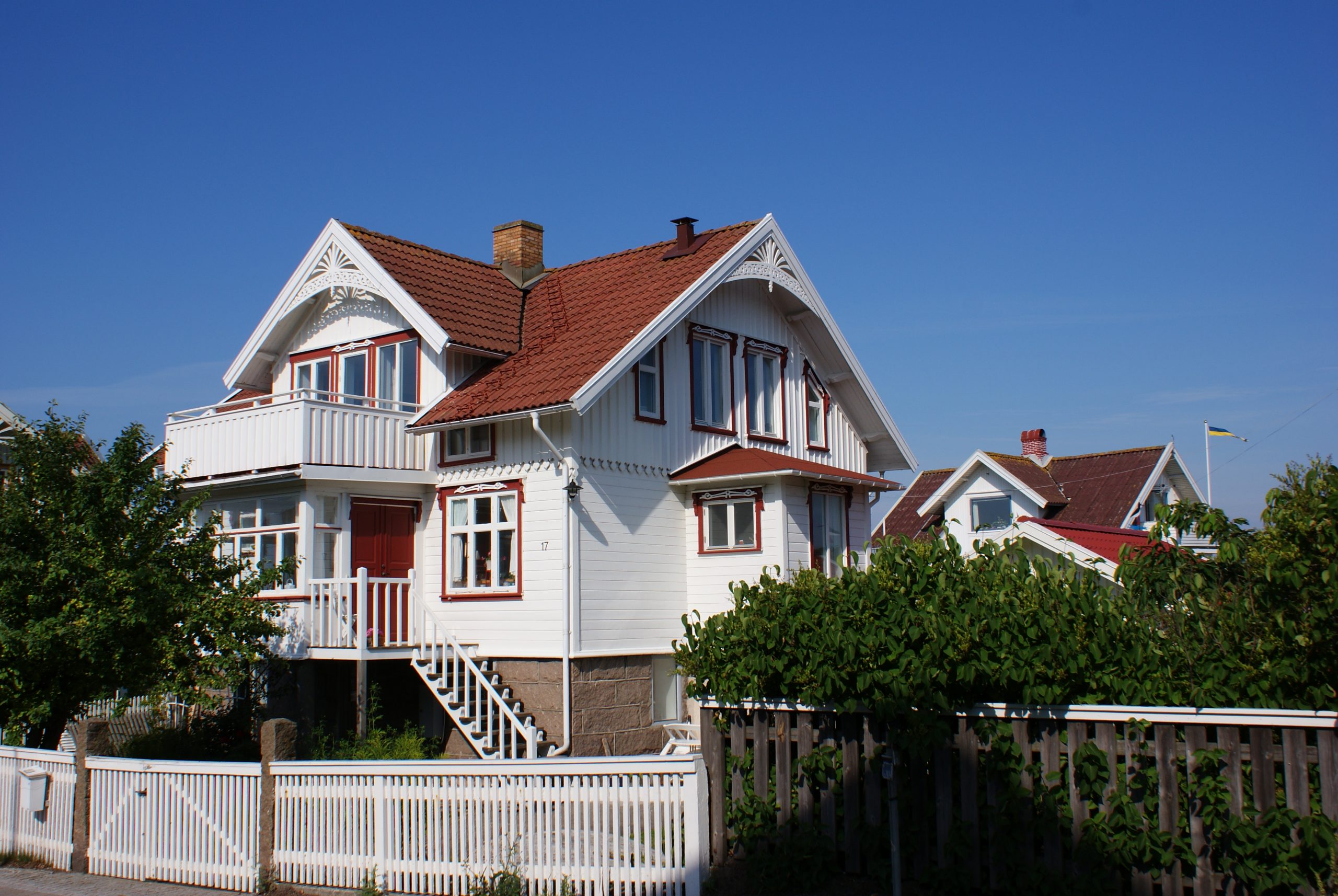 Foto: Ljusmålat bostadhus med veranda, frontespis, röda fönsterfoder och rött tegeltak.