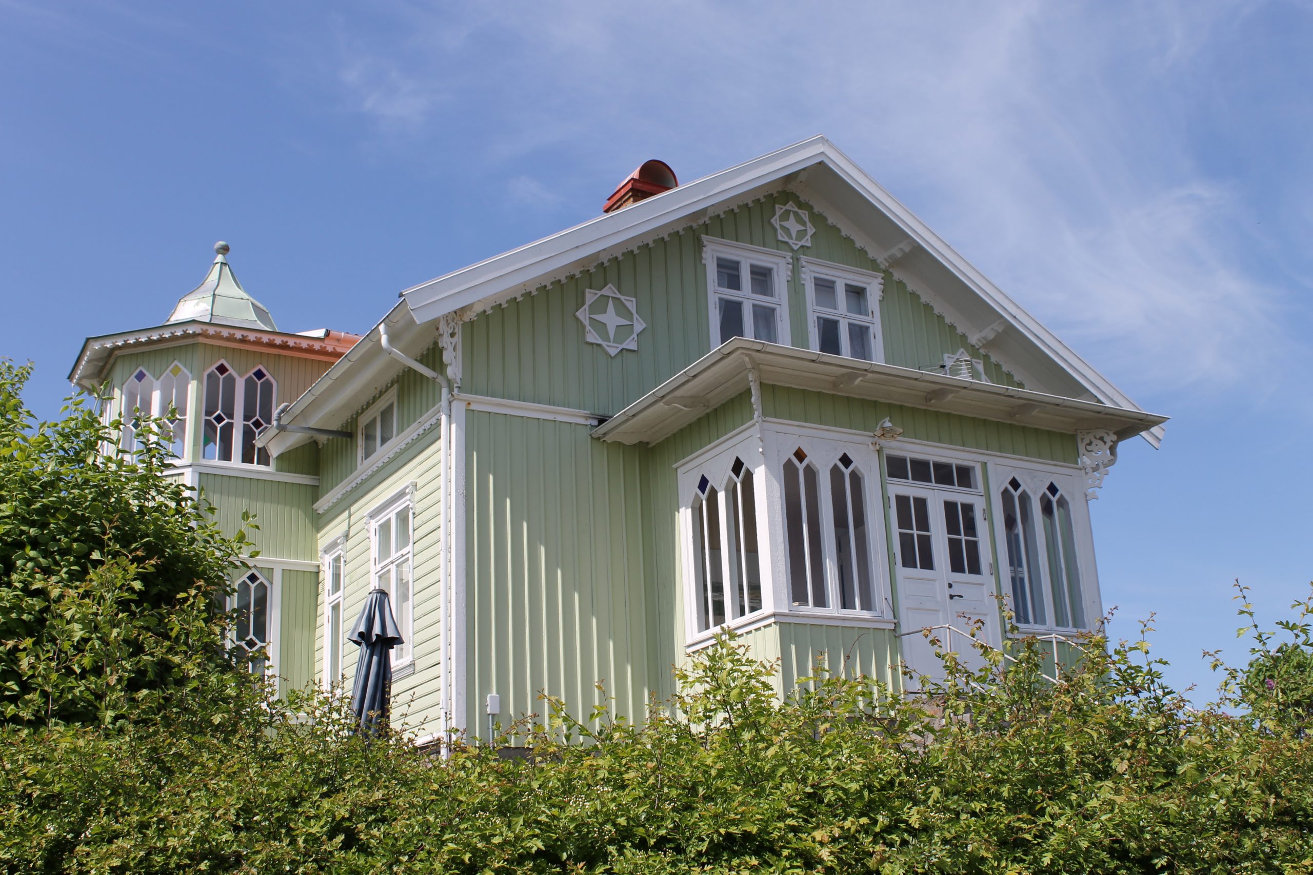 Foto. Ljust grönmålat bostadhus med vita snickerier. Utbyggd veranda och trapphustorn med spröjsade fönster.