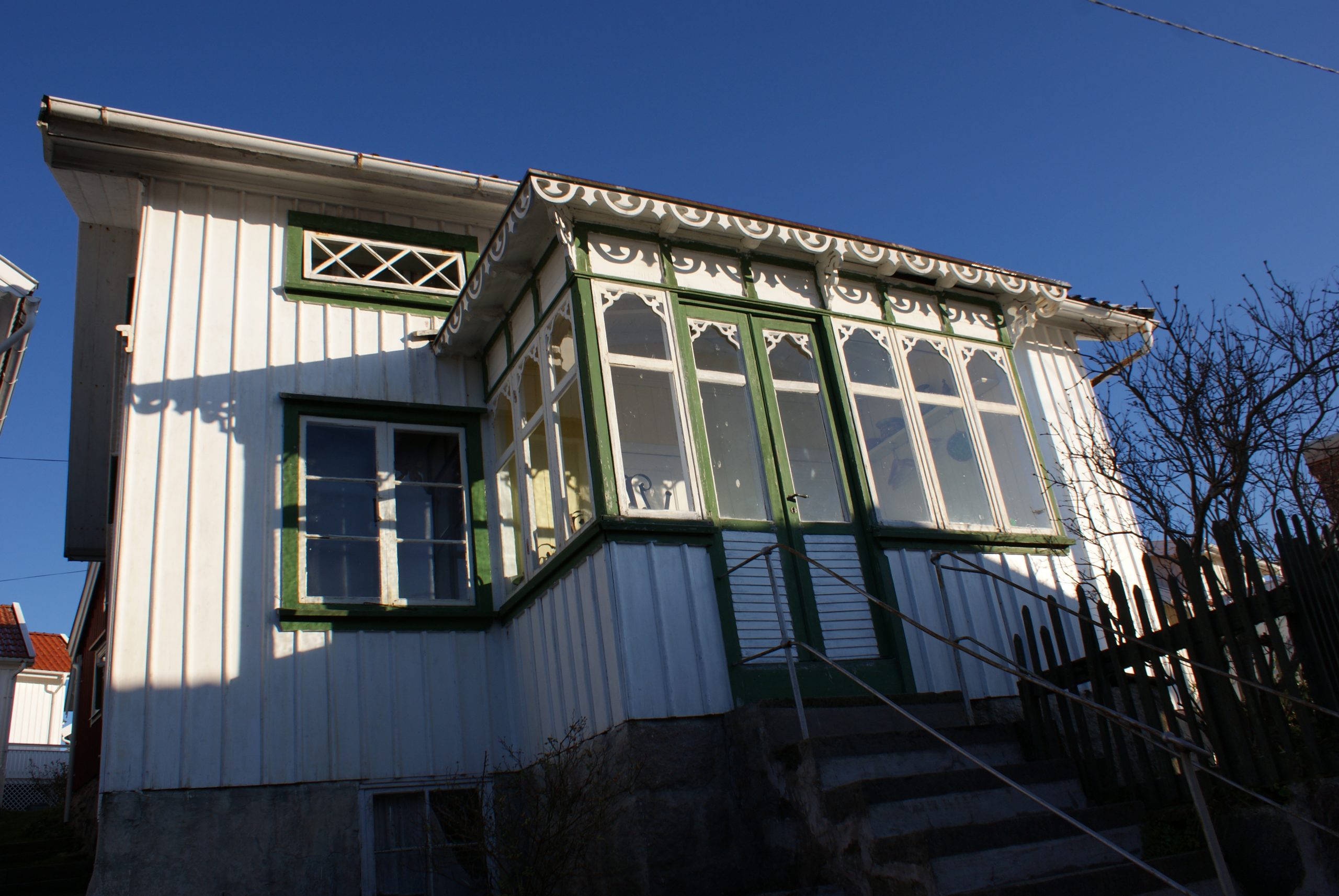 Foto. Ljusmålat bostadhus med utbyggd veranda. Gröna foder och vita snickeridetaljer.