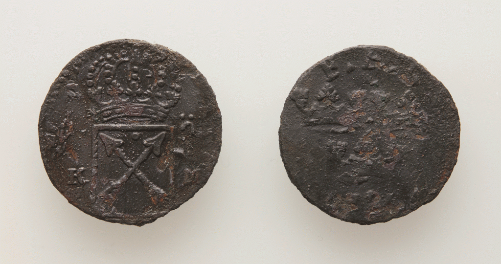 Två mörkbruna cirkelrunda mynt med präglat mönster och enstaka rostfläckar. På det vänstra är konturerna av en krona och under den en rektangel med två korslagds spjut. På den högra går inte mönstret att urskilja.