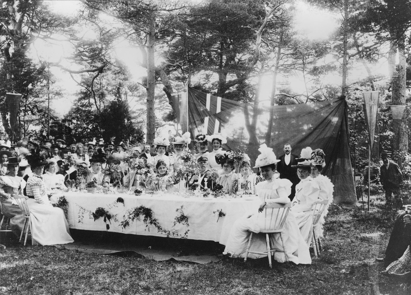 Svartvitt foto. Ett tiotal festklädda damer med kungen i centrum, sitter utomhus runt ett dukat bord. Runt om står en stor grupp damer och herrar.