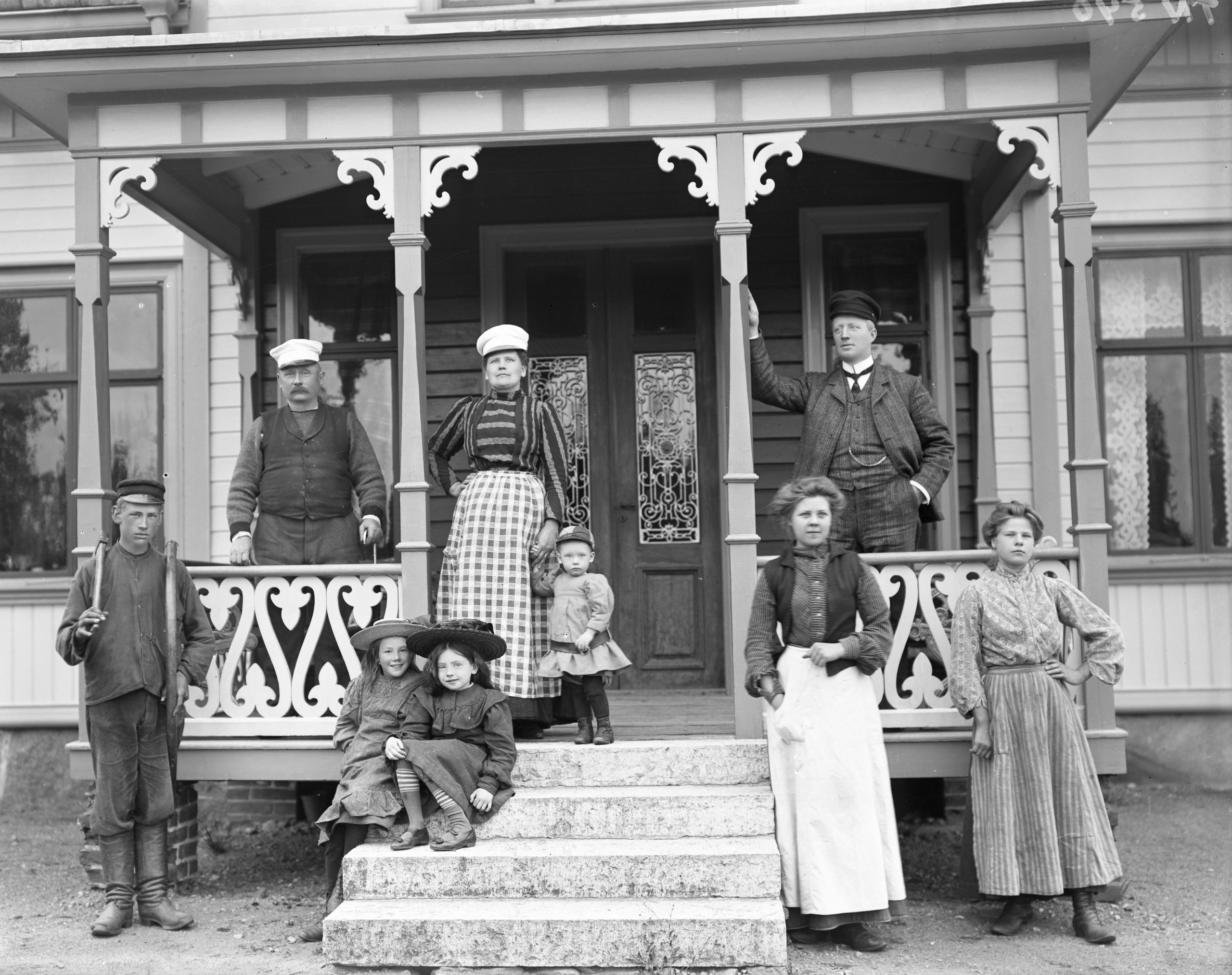Svartvitt foto. Gruppbild med tjänstefolk i arbetskläder vid en veranda, tre män, tre kvinnor och tre barn.