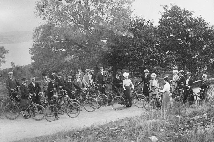Svartvitt foto. Gruppbild med tretton manliga och åtta kvinnliga cyklister som står på vägen med sina cyklar.