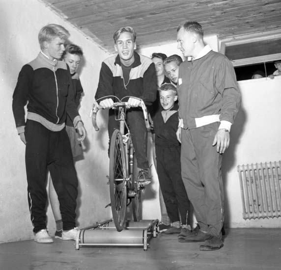 Svartvitt foto. En ung man på en cykelmasin omgiven av fem ynglingar och en tränare alla klädda i träningsoverall.