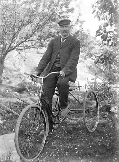 Svartvitt foto. Man i kostym och keps på en stor trehjulig cykel.