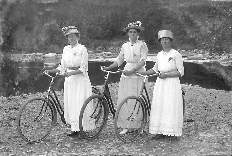 Svartvitt foto. Tre damer i hattar och ljusa långa dräkter står vid sina cyklar vid stranden.