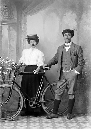 Svartvitt ateljéfoto. En kvinna i lång kjol och hatt står med en cykel bredvid en man i sportkostym. 