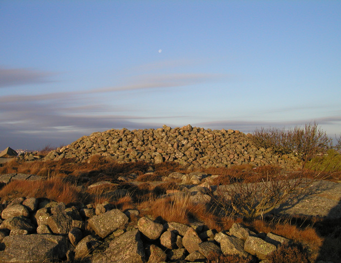 Foto. Ett röse av rundade stenar som är 10 meter i diameter och 1 meter högt. På den ljusblå himlen ovanför syns en blek halvmåne och tunna molnskyar.