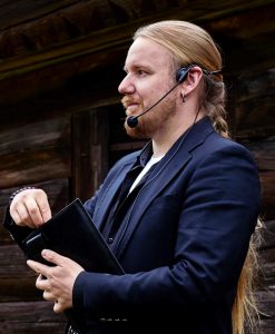 Foto. Tommy Kuusela i profil. Håller ett föredrag, har håret i en lång fläta, mörkblå kostym och mikrofon headset på sig.