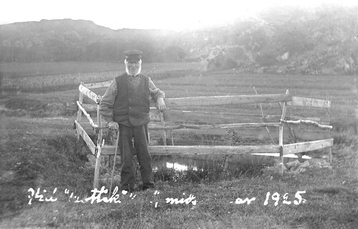 Svartvitt foto. En äldre man med väst och keps står vid en källa inhägnad med ett trästaket.