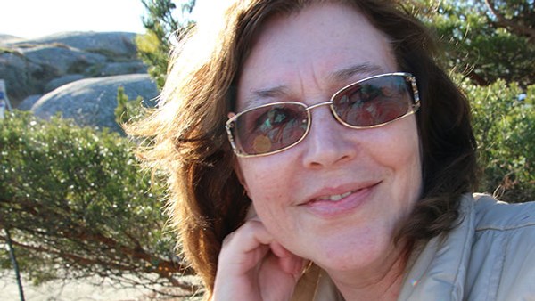 Porträttfoto på föredragshållare. Kvinna, brunt hår, solglasögon. Ler och tittar i kameran.