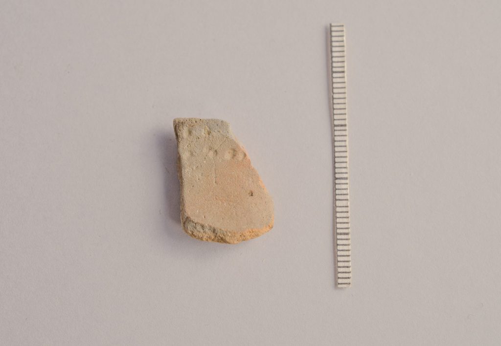 Foto. En lerskärva med små runda intryckta punkter. En skalstock visar att skärvan är ungefär 10 centimeter stor.