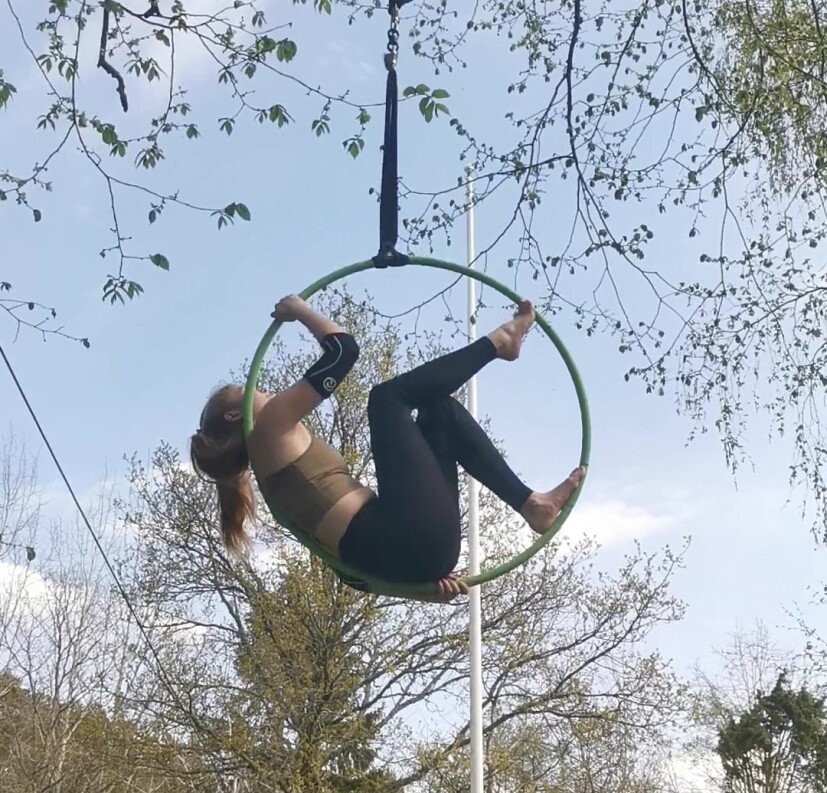 Foto. En kvinnlig akrobat hänger inuti en ring fastsatt i ett träd.