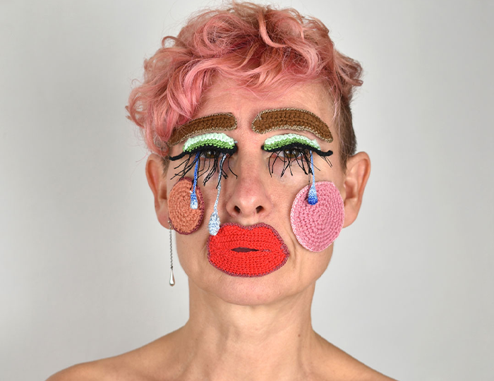 Photo. A close-up shot portrait of a woman with short, pink hair. On her face lays crocheted pieces depicting makeup in bright colours.