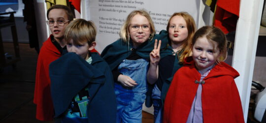 Foto: Fem barn i tioårsåldern tittar in i kameran. De bär mantlar i färgerna rött och blått.
