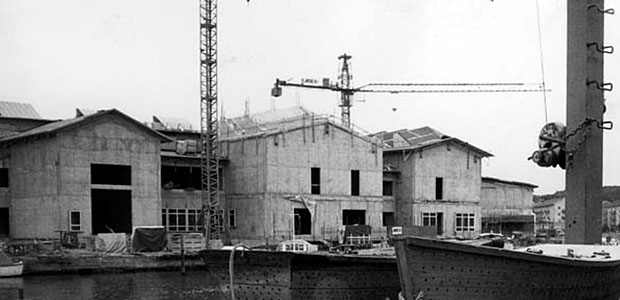 Svartvitt foto av pågående husbygge, med tre ofärdiga byggnadskroppar och två lyftkranar.