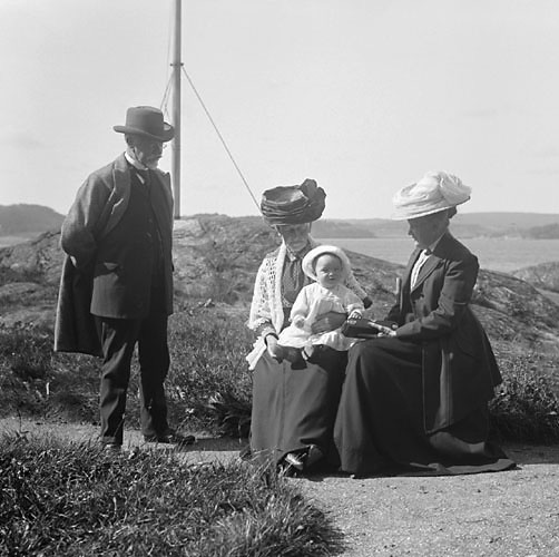Svartvitt foto. En äldre man står och två älder damer i stora hattar sitter med liten baby vid en promenadväg.