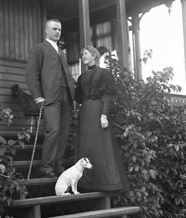 Svartvitt foto. En ungt par i mörka dräkter står tillsammans med en liten hund på en verandatrappa.