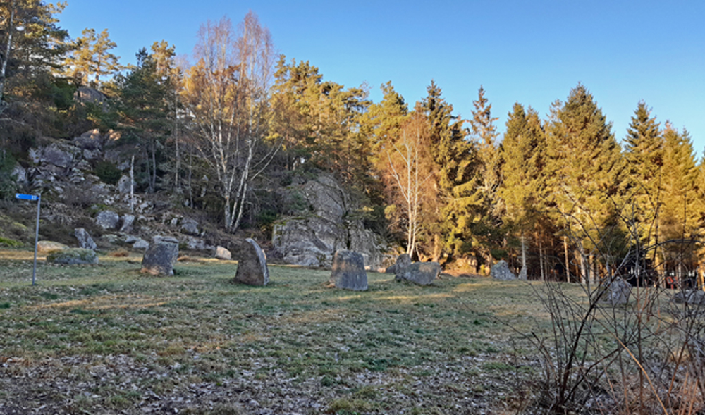 Färgfoto av gräsyta framför berg. På ytan står ett antal knappt meterhöga stenar i cirklar. 