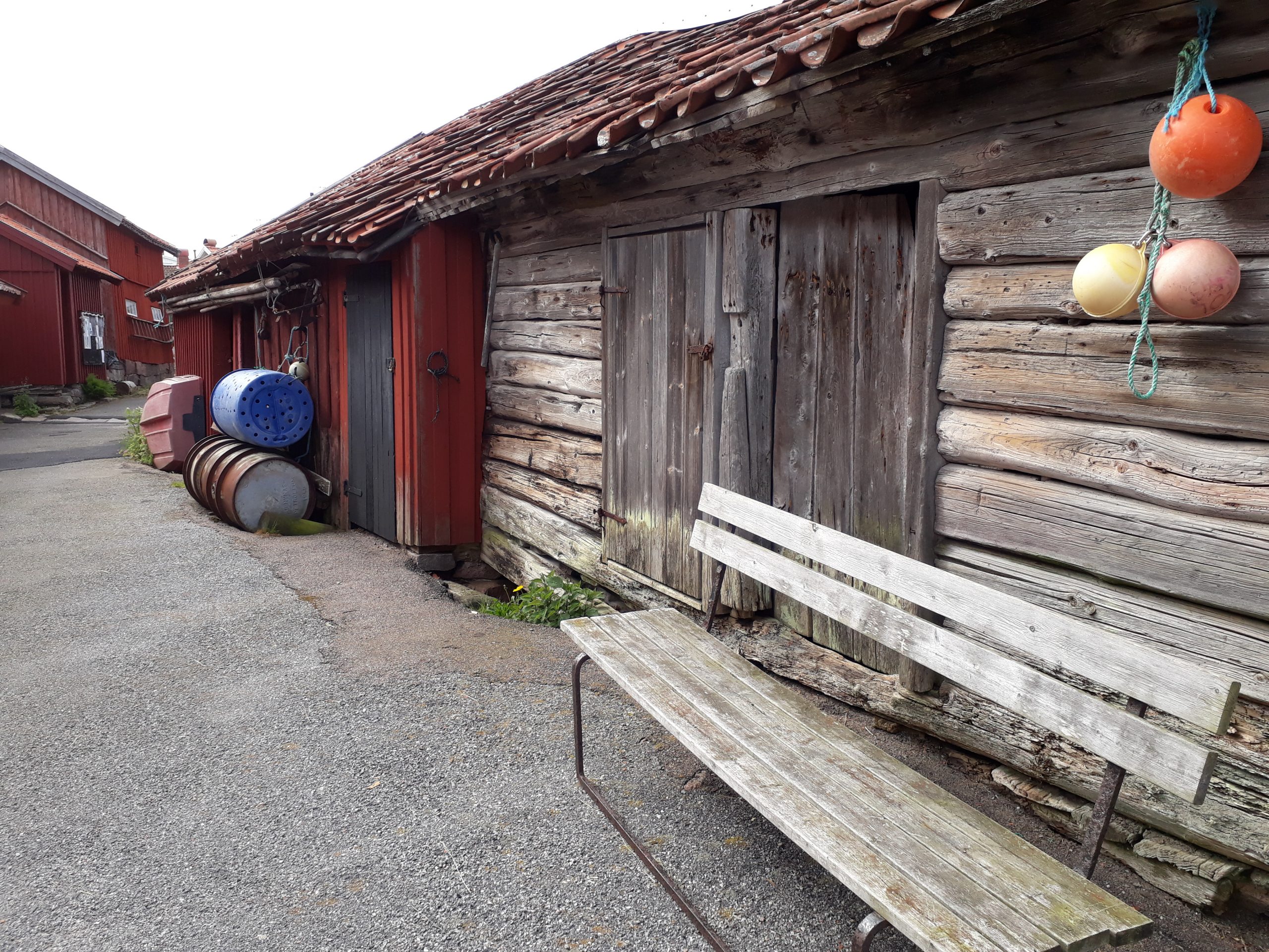 En grå sliten träbänk står framför en gammal omålad sjöbod. bakom den står en rödmålad sjöbod. Bodarna står invid en gångväg som leder bortåt.