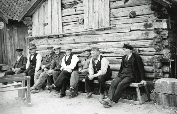 Svartvitt foto sex män som sitter på en äldre träbänk framför en omålad sjöbod.