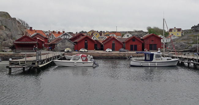 Färgfoto på en stilla hamn med två båtar i vattnet. Bakom bryggan och hamnplanen ligger en rad med sjöbodar. Mellan de två längst till vänster skymtar gångvägen upp till samhället.