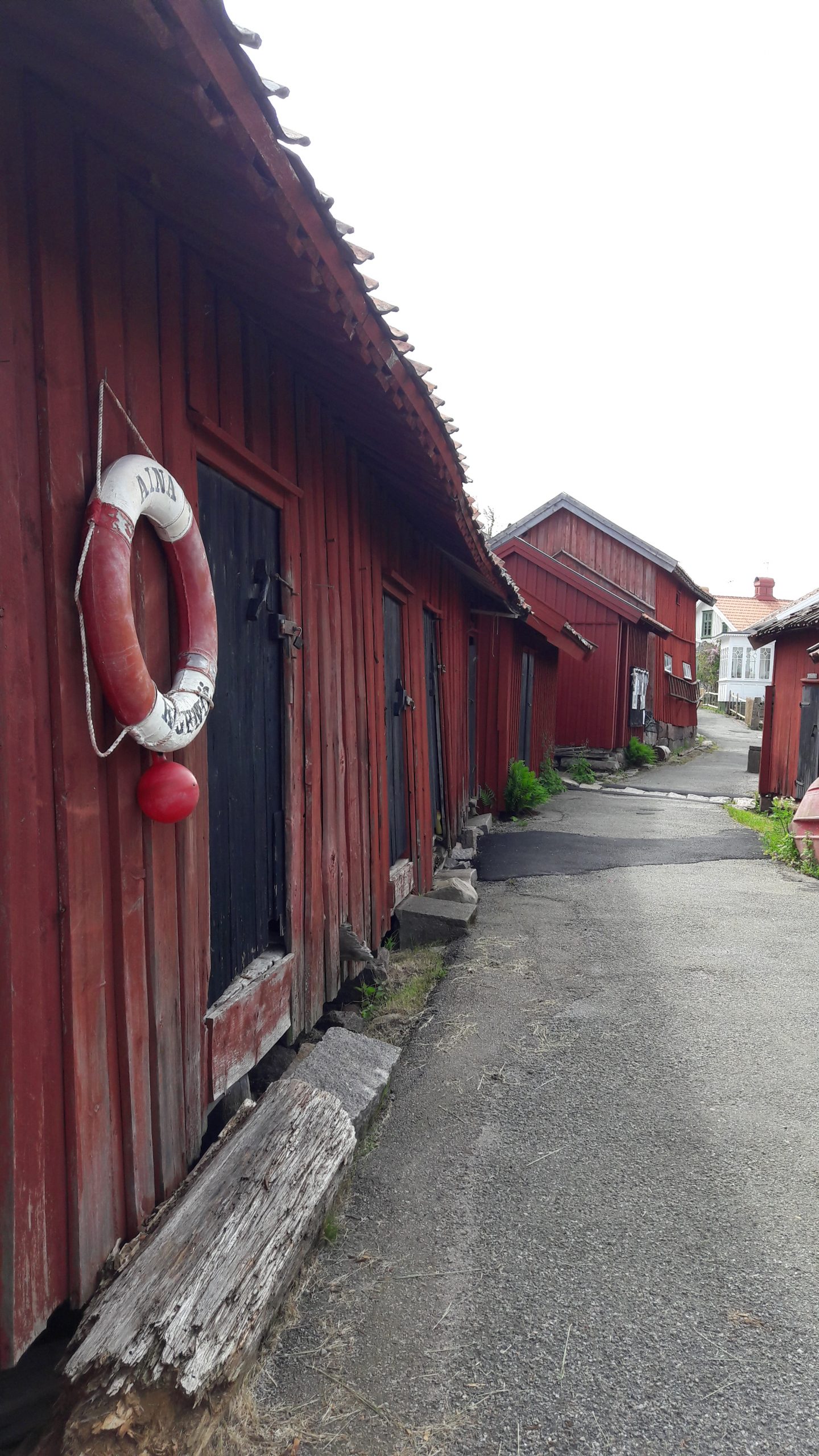 Färgfoto med röda sjäbodar till på ömse sidor en smal asfalterad väg som leder in till samhället.