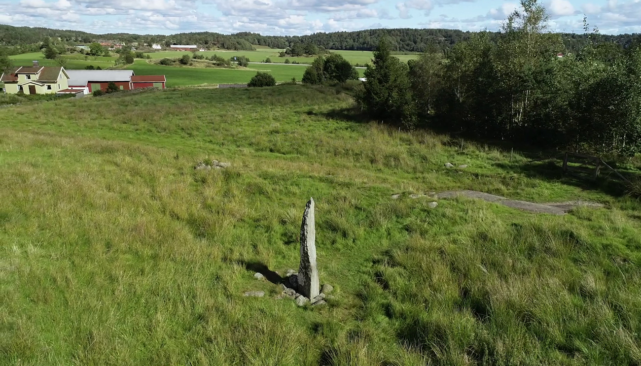 Färgfoto med en gräsklädd kulle med utsikt bortåt i fjärran. Bakom kullen ligger några hus och mitt på kullen står runstenen.