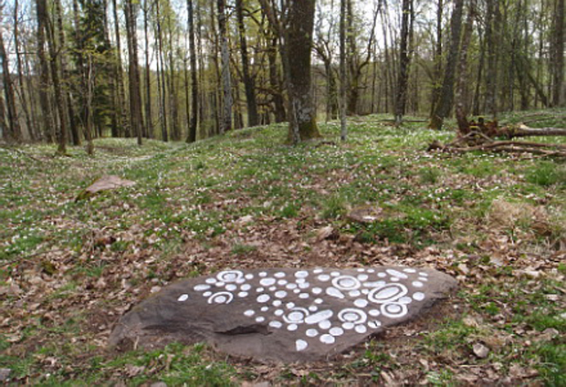 Färgfoto av ett stenblock med vitmålade figurer som ligger i en gräsklädd hage med vitsippor och lövträd.