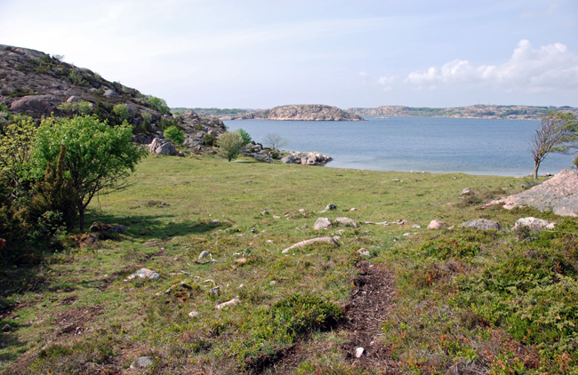 Grässlänt som sluttar ner till havet. På ömse sidor slänten sticker det upp berg och ute i havet ses flera grå öar.