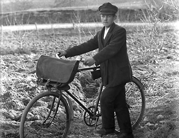 Svartvitt foto av en yngling i keps vid sin cykel med väska på styret.