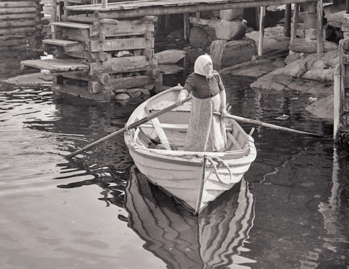 Svartvitt foto av en kvinna med huckle, stående i en roddbåt.