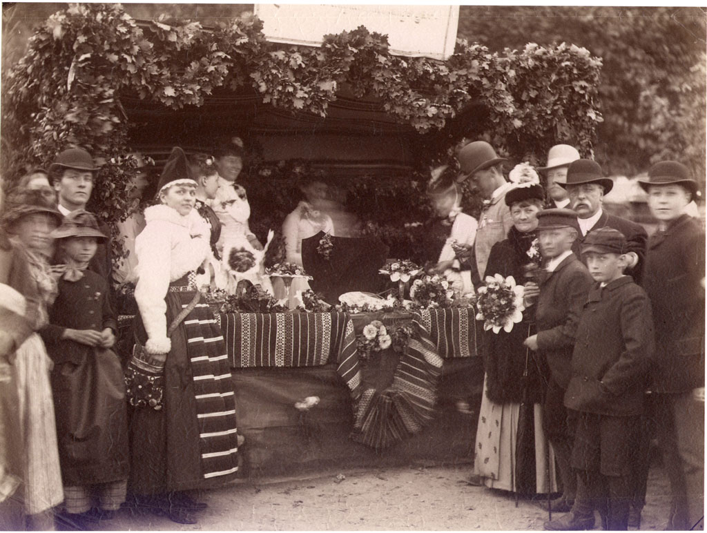 Människor vid ett marknadsstånd med blomsterarrangemang. En kvinna klädd i folkdräkt.