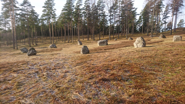 Foto. I en grässlänt står 11 meterhöga stenar i en cirkel, cirka 15 meter i diameter.