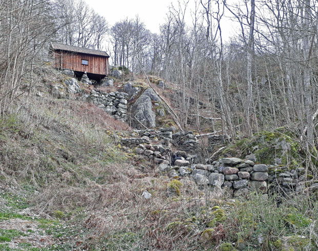 Foto. Kvarnbyggnaden, en liten brun träbyggnad, står på stenstolpar ovanför en brant med sten-ruiner.