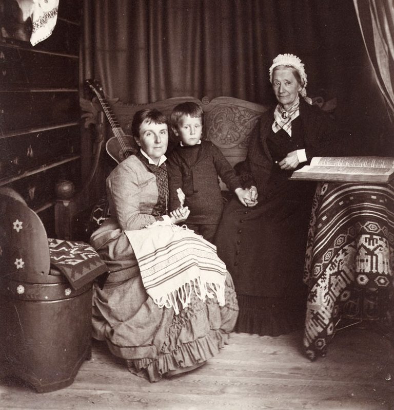 Svaratvitt foto. Två r sittande kvinnor i långa klänningar håller en liten pojke i händerna.