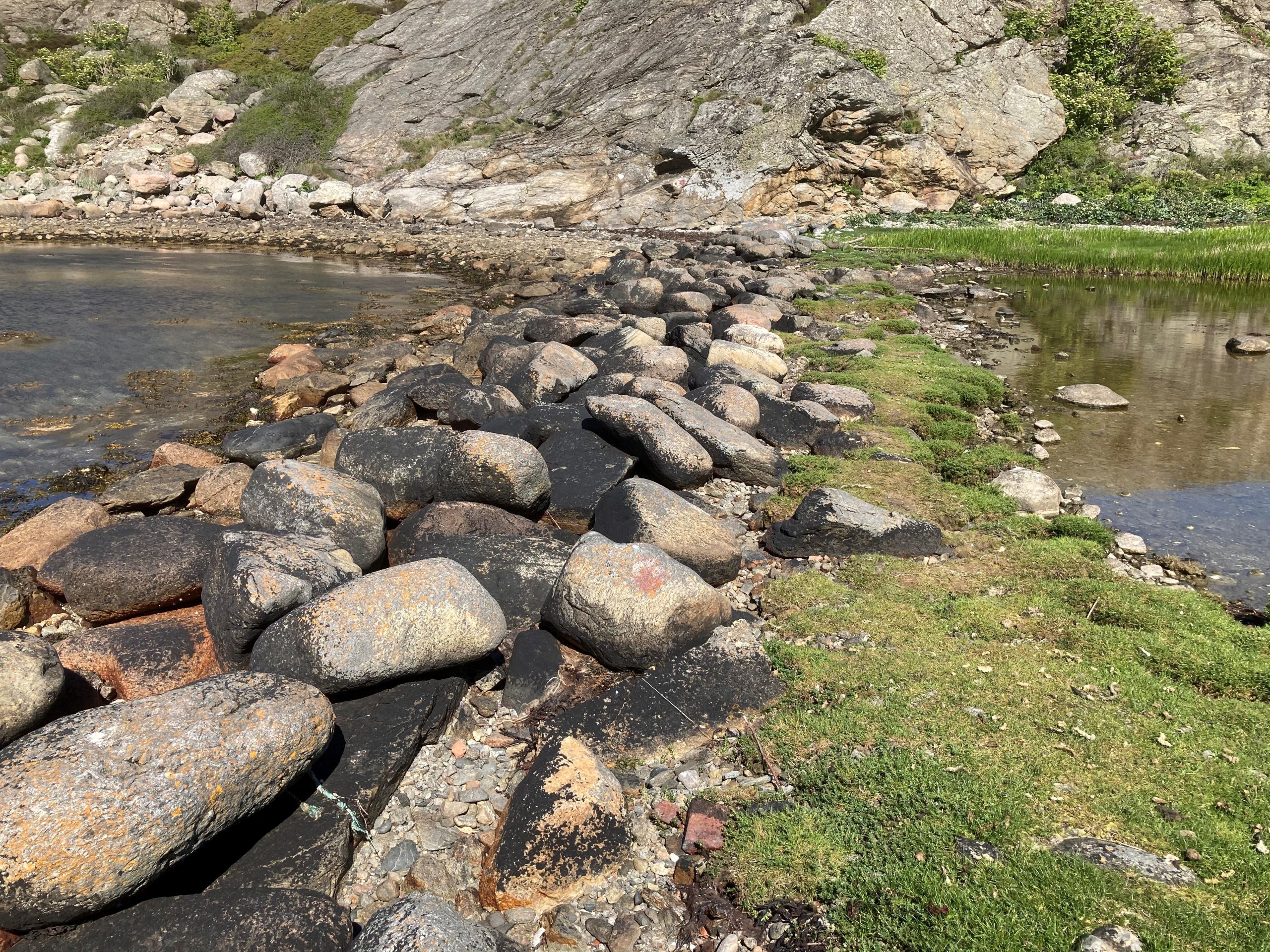 Färgfoto av stenmur tvärs över en vik. De stora gråsvarta stenarna är närmast havet och innan för en gräsbetäckt yta. 