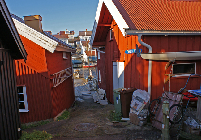Färgfoto på rödmålade bodar. Emellan bodarna skymtar bryggan och vattnet i det smala utrymmet.