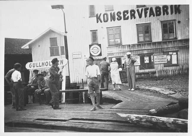 Svartvitt foto. Fem män med keps samtalar på en brygga. Bryggan angränsar till en träbyggnad, en konservfabrik.
