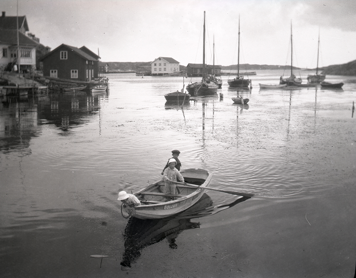 Svartvitt foto av barn i eka i vatten. I bakgrunden flera mindre segelskutor av äldre typ.