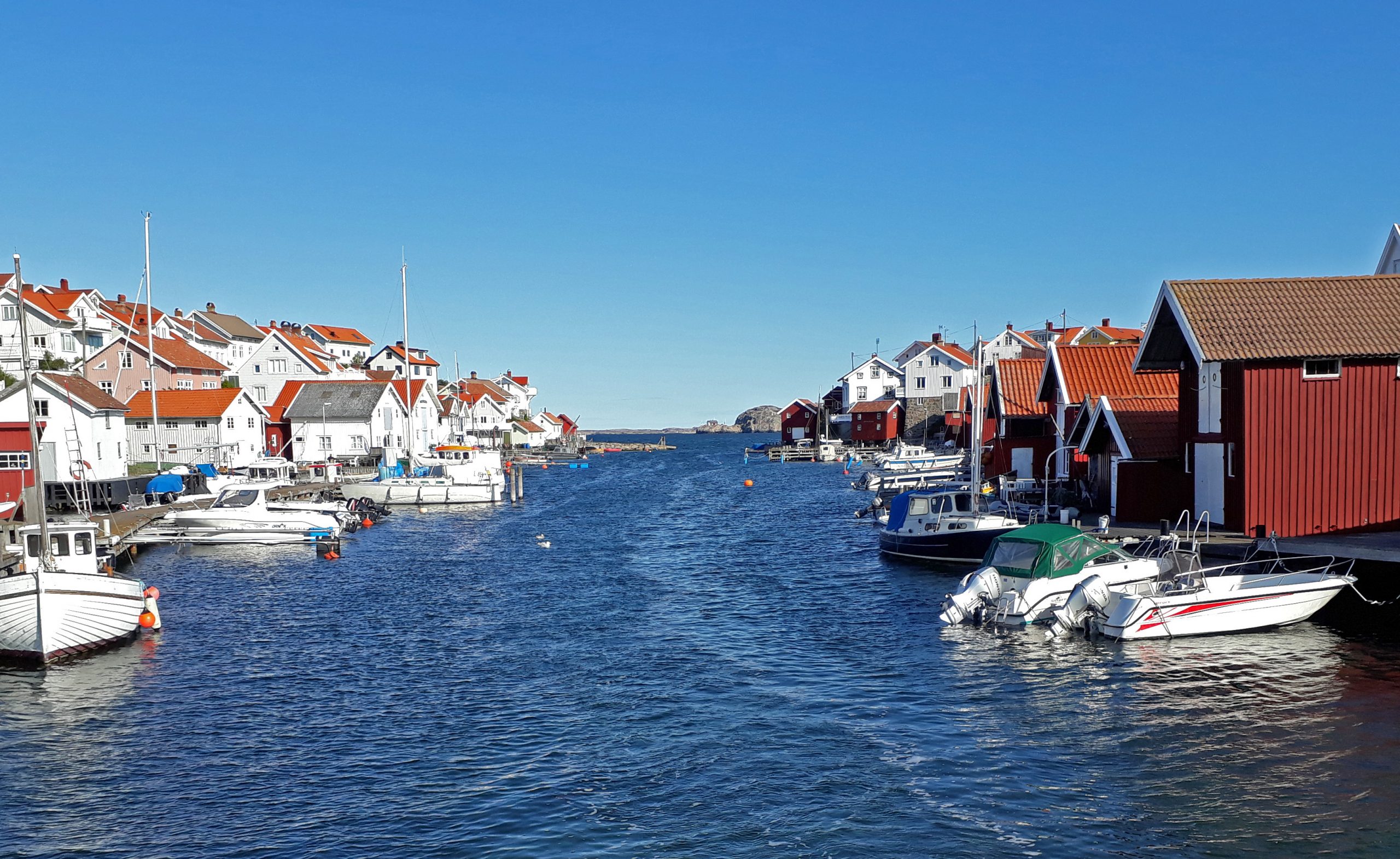 Färgfoto av blått vatten i smalt sund med båtar, bryggor och sjöbodar på sidorna.