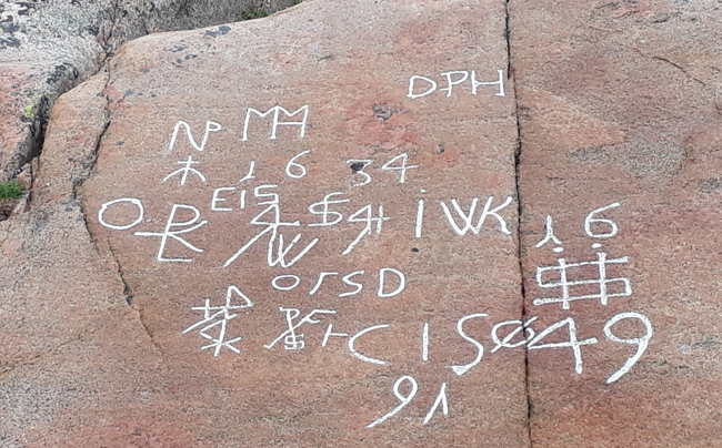 Färgfoto av gråbrun berghäll med ristningar imålade med vit färg.