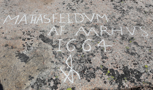 Färgfoto på grå berghäll med inristat namn, bomärke och årtal imålade med vit färg.