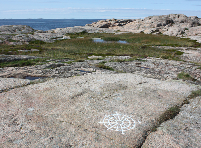 Färgfoto på berghällar, en vattensamling med gräs runt och öppet blått hav i bakgrunden.