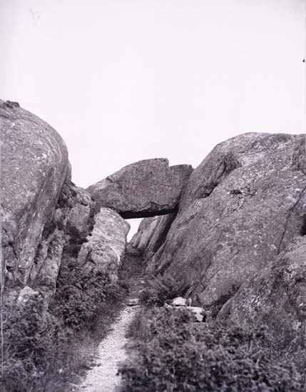 Svartvitt foto. Ett stor klippblock vilar på bergssidorna över en smal gånväg.